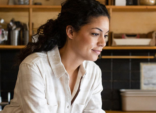 A woman is working on a laptop at the kitchen table, which has a notepad and a pen, a stack of papers, and two slices of cake on a wooden board.